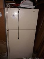 Front view of cream/off-white two-door refrigerator with plug hanging in front. Top freezer and bottom fridge compartments visible.