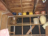 Shelving in a barn or garage showing multiple ceramic pots and crocks, including a brown spittoon and beige crocks.