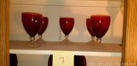 Five red glass wine goblets on a shelf with a wooden frame, clear stems and bases, measurements noted behind for scale.
