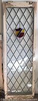 Full view of antique door with leaded glass and stained glass shield recessed in diamond pane pattern. Door shows worn beige paint and two hinges at top.