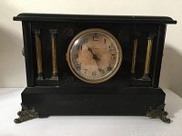 Front view of an antique black mantle clock with a circular dial and decorative columns on each side of the face. Clock face shows aged dial with faded numbers and black hands.