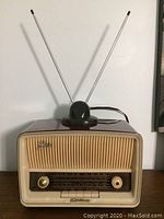 Front view showing vintage radio with ribbed cream grille, gold control knobs, and twin-rod antenna extended.