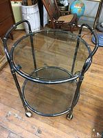 Full view of the vintage metal bar cart showing its round glass shelves and wheels on a wood floor.