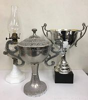Front view showing all three items: the tall shiny silver trophy, the speckled trophy with lid, and the white based oil lamp.