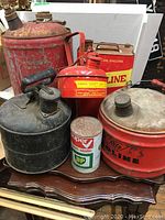 Overview photo of the lot showing five various vintage metal gasoline cans in red and black with visible wear and rust.