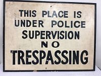 Front view of vintage metal sign with black text on a cream background, displaying 'This place is under police supervision No trespassing'. Shows marks and wear.