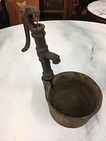 Miniature metal water pump standing on a white surface showing rusted detailed body and bowl base.