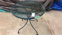 Front angle view of the green metal patio table with mesh top and tripod legs on concrete floor with covers in the background.