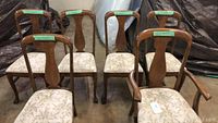 Group view of all six dining chairs, five armless and one with wooden armrests, showing quarter sawn oak wood backs and leaf patterned upholstered seats.