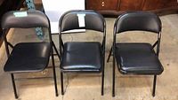 Three black vinyl folding chairs shown side-by-side, seats and backrests made of vinyl, metal frames painted black.