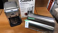 Photo of Cuisinart coffeemaker with box and stainless steel breadbox on wooden table.
