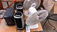 Two white table fans, two Bionaire tower fans, and one Dell black speaker on a wooden surface
