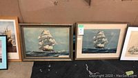 Several framed ship artworks leaning against a wall showing various sailing ships on the ocean with different frames and art styles.