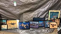 Wide shot showing all 14 glossy airplane art prints arranged horizontally on a floor with a fabric backdrop, displaying various WWII aircraft in flight and airshow scenes.