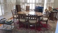 Dining table set including table and six chairs shown fully arranged on red patterned rug near large windows