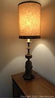 Full view of lit lamp showing cylindrical linen shade and multi-tiered bronze base