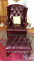Front view of burgundy leather wingback chair with button tufting, matching ottoman and throw pillow