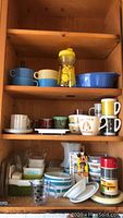 Shelf of assorted vintage Pyrex bowls, mugs, and dishes shown including blue geometric patterned bowls, yellow handled sippy cup, and various colorful mugs with mid-century designs