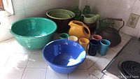 Overview of vintage green glass kitchen accessories, ceramic mixing bowls, Fiesta Ware pitcher and tumblers, and copper tea kettle on countertop.