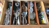 Drawer with compartments holding various silverware including spoons, forks, knives, and small glass dishes.