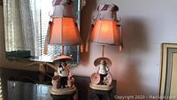 Pair of lamps with ceramic Chinese figurines shown on black surface, each with lampshade and hooks for jewelry display.