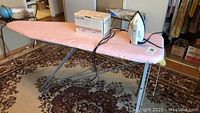 Photo showing the pink padded ironing board with metal frame set up, on a carpeted floor with a white iron placed on top.