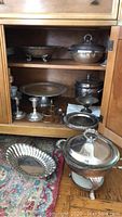 Cabinet shelf and floor with multiple silver plated and sterling items including compote, lidded ice bucket, plates, and candleholders.