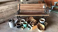 Front view of wrought iron and wooden garden bench with assorted ceramic pots, wicker baskets, and metal watering can arranged on ground in front