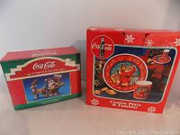 Photo showing the original boxes for both Coca Cola Santa collectibles: the Santa in Sleigh figurine box and the cookie plate with tumbler set box.