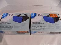 Photo of two new boxed virtual reality smartphone headsets, blue and black, showing front and side of boxes with branding and product image.