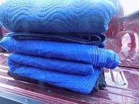 Five folded moving blankets stacked on back of red pickup truck bed, showing blue and black colors and quilted pattern.