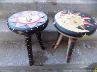 Two 15-inch tall hand painted three-legged stools, one with a sun design, the other with moon and star design on the tops, placed on concrete steps.