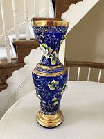 Full view of tall blue glass vase decorated with hand painted white and yellow flowers, gold scrollwork, and gold painted top rim and base.