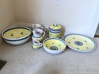 Full view of all seven ceramic pieces arranged on a carpet against a wall. Includes bowls, pitchers, lidded jar, and serving plates with visible colors and designs.