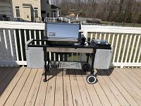 Front view of black metal outdoor charcoal grill with silver lid, temperature gauge, two large side shelves and wheels, placed on deck.