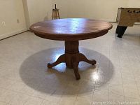 Side view of a round wooden dining table with pedestal base on a tiled floor, showing overall shape and base design.