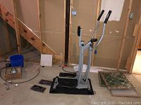 Exercise strider machine positioned in unfinished basement with visible wooden staircase and wall framing.