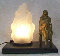 Full view of the lamp showing the lit flame-shaped shade and the seated lady sculpture on a rectangular black base.