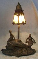 Front view of the gilt metal lamp showing two nude mermaid figures on a dolphin base with caramel stained glass shade lit.