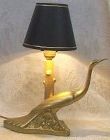 Front perspective view of the gilt metal Art Deco bird table lamp illuminated, showing the bird figure attached to the triangular base and the black shade with gold trim.