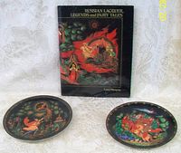 Two Russian fairy tale decorative plates with bright traditional scenes and the illustrated hardcover book, all set on a white textured fabric.