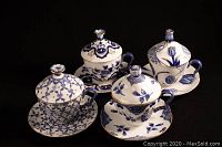 Four blue and white china covered cups with matching saucers arranged in square on a black background. Each cup has a lid with a flower-shaped knob and distinct blue floral pattern.