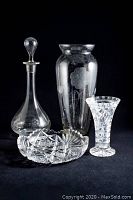 Clear etched and cut crystal decanter with stopper, etched vase, cut crystal bowl, and a 6-inch cut crystal vase arranged together on black background.