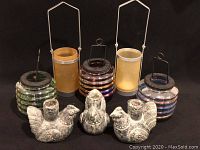Photo showing four outdoor garden lanterns with metal handles and ribbed colored glass. In front are three vintage stoneware hen candleholders in gray tones with worn texture.
