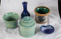Five handmade pottery pieces placed on white cloth, showing the signed porcelain jar in pale turquoise glaze alongside other pottery including a blue vase, green pot, beige pot with brush strokes, and small blue dish.