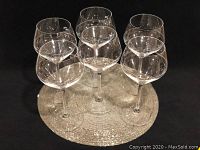 Six clear etched crystal balloon wine glasses arranged on a decorative silver placemat against a black background.