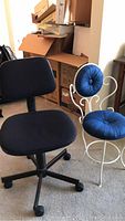 Photo showing black fabric desk chair with swivel base and small white iron bistro chair with blue cushions placed side by side in a room with boxes and a table visible in the background.