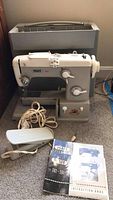 Front view of Pfaff model 362 sewing machine with attached cords and footswitch, placed in front of the carrying case. Manuals placed nearby.