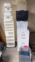 Full lot showing white metal double drawer file cabinet on casters, tall clear plastic rolling drawer unit, stacked plastic containers, metal mesh file cube, black metal lockable box, and metal storage boxes.