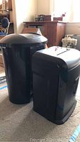 Side view of the black Staples paper shredder next to the Umbra black lidded trash can showing general shape and size comparison.